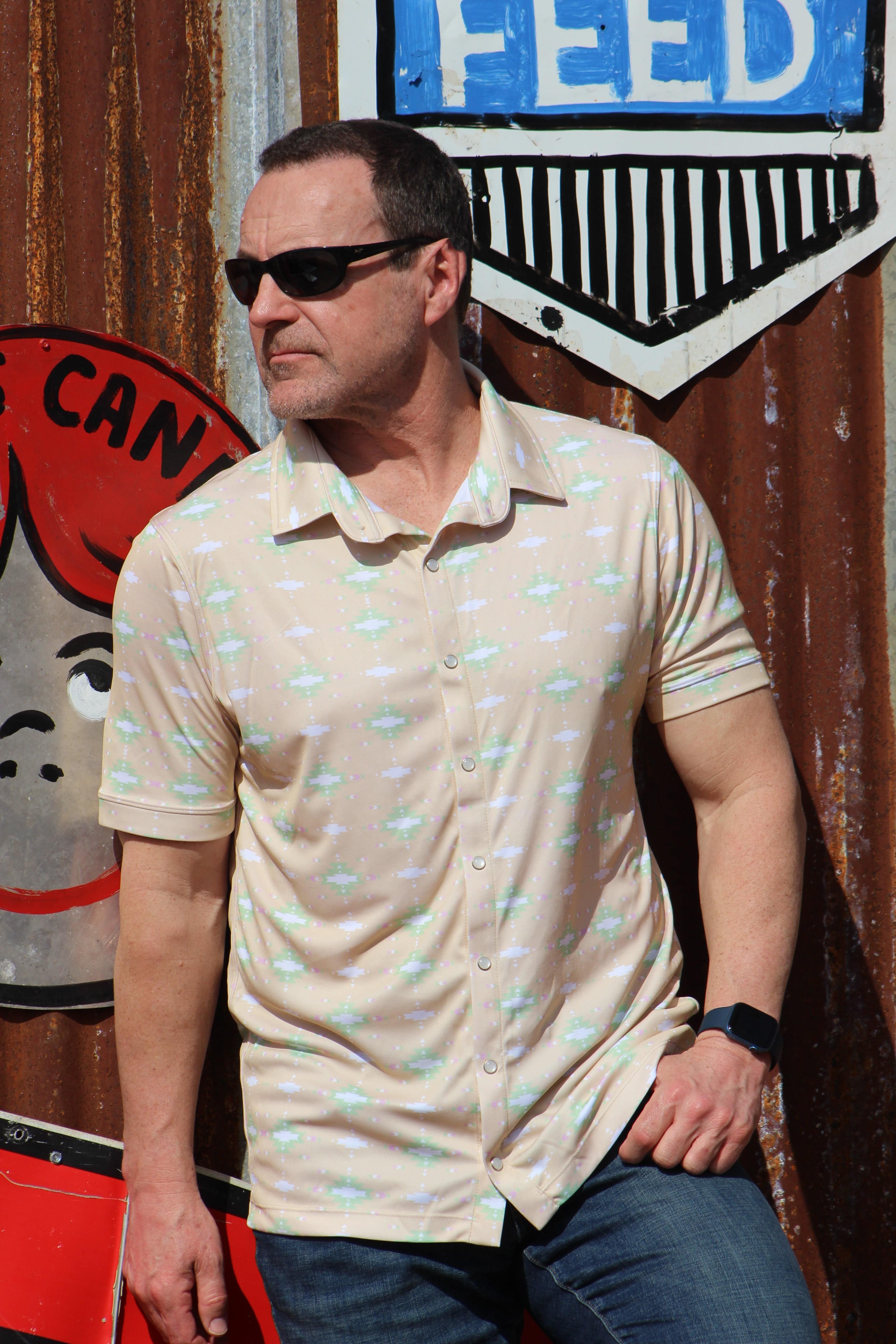 A man exudes cool confidence in HowdyHo's Sunrise Serenity shirt, a perfect blend of casual elegance and comfort. The shirt's soft tan color with a light green and white floral pattern provides a fresh and relaxed look. Accessorized with sleek sunglasses and paired with classic denim jeans, this ensemble is set against a colorful, rustic urban background, showcasing the shirt's versatility for any laid-back occasion.