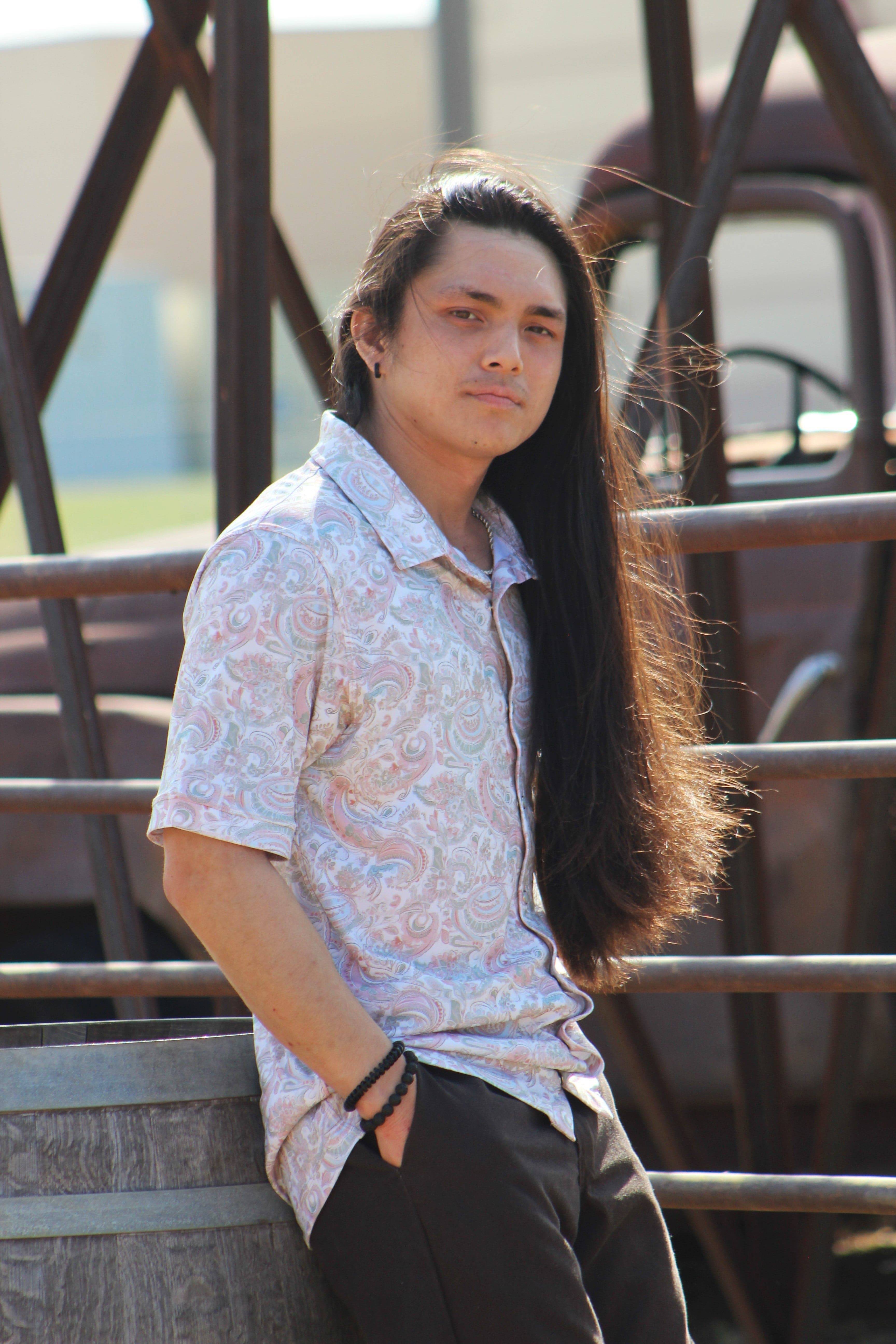 A man leans casually against a metal structure, modeling the 'Green Love Paisley' shirt available on Howdy-Ho.com. The shirt features a delicate paisley print in soft pink and green hues, creating a vintage vibe. He completes his look with dark pants and a black beaded bracelet, embodying effortless style.