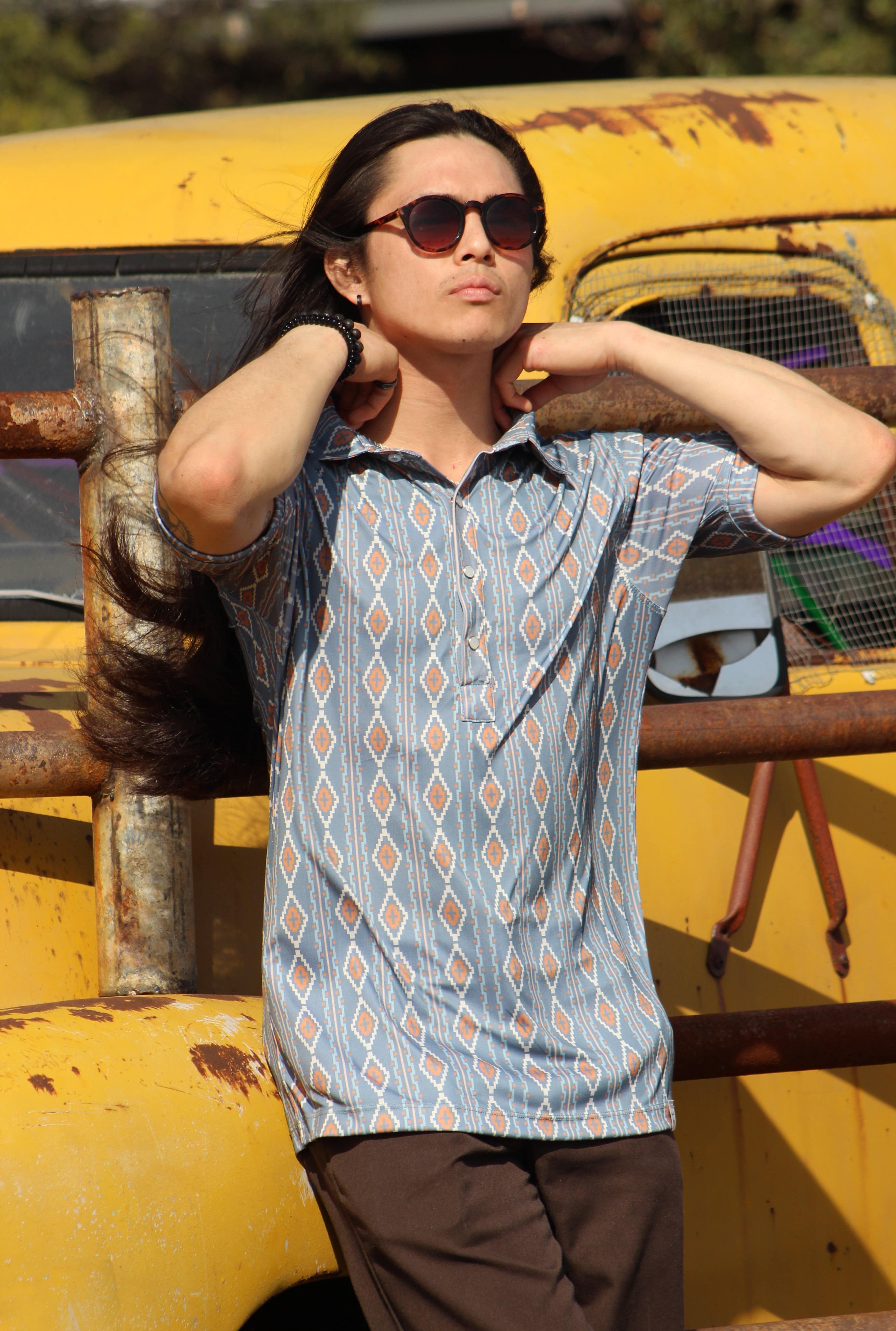 A person models the HowdyHo Aztec Brown Blue shirt with confidence and style. The shirt's striking pattern of geometric shapes in hues of blue and brown adds a bold touch to the ensemble, paired with smart brown trousers. They stand before an aged yellow vehicle, with sunglasses adding a chic finish to the adventurous, masculine aesthetic of HowdyHo's men's collection.