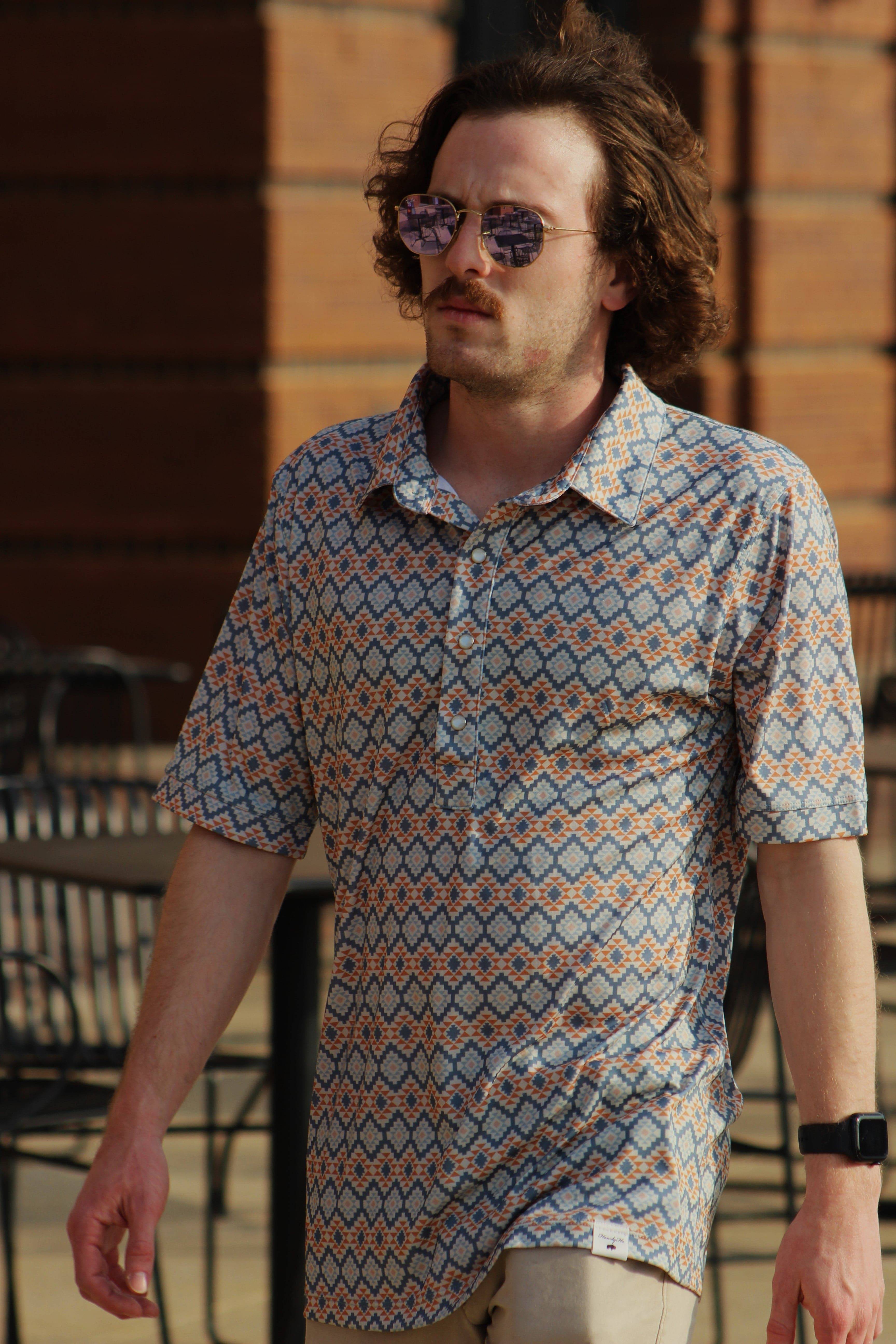 A man walks confidently in an urban setting, wearing the Aztec Twilight Blend shirt from HowdyHo. This stylish piece features a unique blue and orange Aztec pattern on a crisp, button-down shirt with a pointed collar. The shirt's contemporary design is complemented by his cool round sunglasses, adding a modern twist to a classic look.